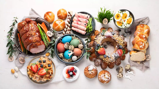 A festive Easter spread with a variety of dishes including glazed ham, deviled eggs, a bundt cake decorated with berries, waffles, and colorful dyed eggs, all arranged on a white tablecloth with small decorative accents.