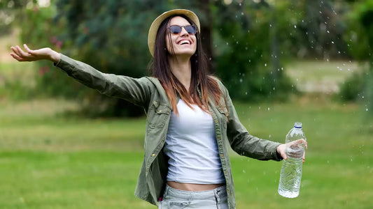 A smiling, eco-friendly woman wearing a hat and sunglasses with her arms outstretched, holding a reusable water bottle, enjoying a light mist of water in a green outdoor setting, embodying natural health and wellness.