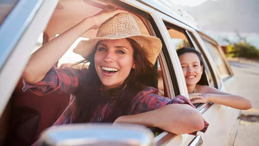 A happy woman wearing a straw hat smiles and looks out of a car window, with a child smiling in the back seat, suggesting enjoyable and carefree road trip travel with healthy eating.